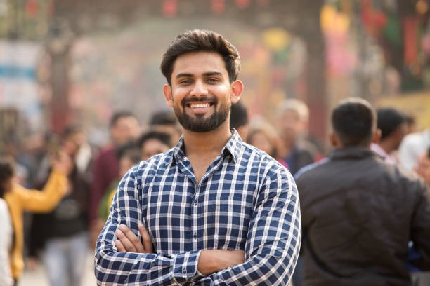 A smiling man with a beard wearing a checked shirt standing confidently with arms crossed in a crowd.